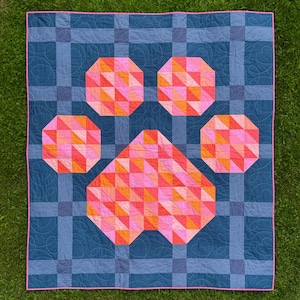 May include: A rectangular quilt featuring a large paw print design composed of pink, orange, and red triangular fabric patches. The background consists of a dark blue and grey plaid pattern with continuous heart-shaped quilting and pink trim.