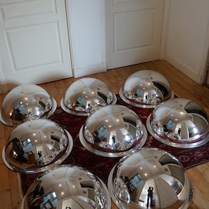 May include: Eight large, polished silver serving domes with wide rims are displayed on a patterned rug and wooden floor. The reflective surfaces of the domes mirror the room's interior. The domes are a striking, eye-catching feature.