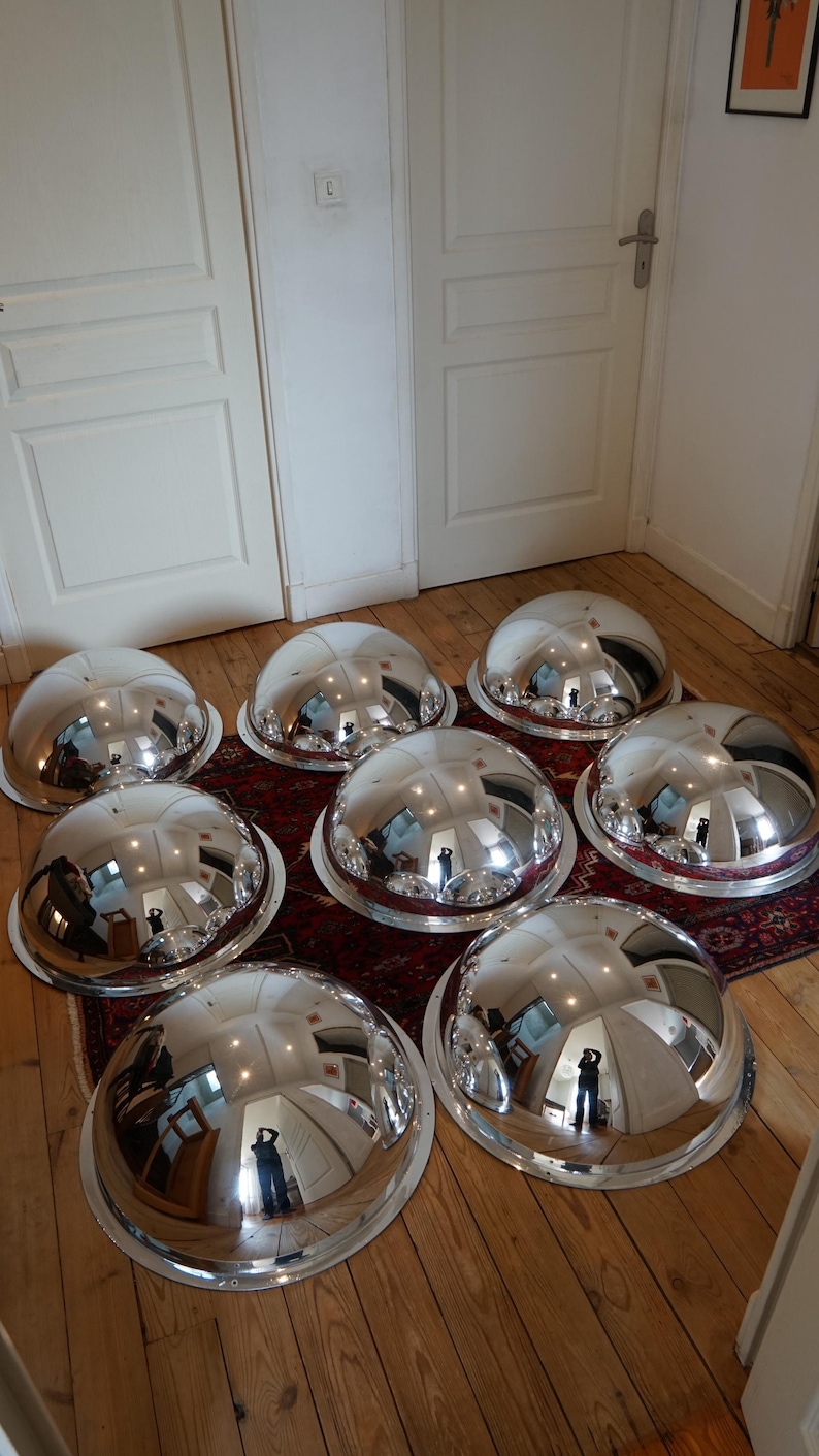 May include: Eight large, polished silver serving domes with wide rims are displayed on a patterned rug and wooden floor. The reflective surfaces of the domes mirror the room's interior. The domes are a striking, eye-catching feature.