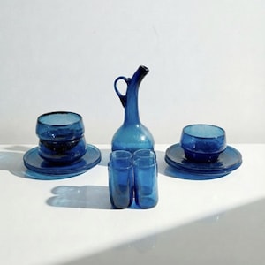 Puede incluir: Un conjunto de vajilla de vidrio azul, que incluye platos, tazas y una jarra con asa. El conjunto también presenta pequeños vasos cilíndricos. Los artículos están dispuestos sobre una superficie blanca, mostrando su transparencia.