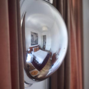 May include: A chrome convex mirror reflecting a bedroom scene. The reflection shows a bed with orange cushions, a white bedside table, and a framed artwork. The mirror is mounted on a wall next to a brown curtain.