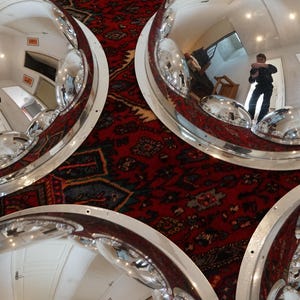 May include: Overhead view of multiple large, polished, chrome-like, hemispherical mirrors with white rims. The mirrors reflect the room and a red patterned rug. The mirrors are arranged in a circular pattern.