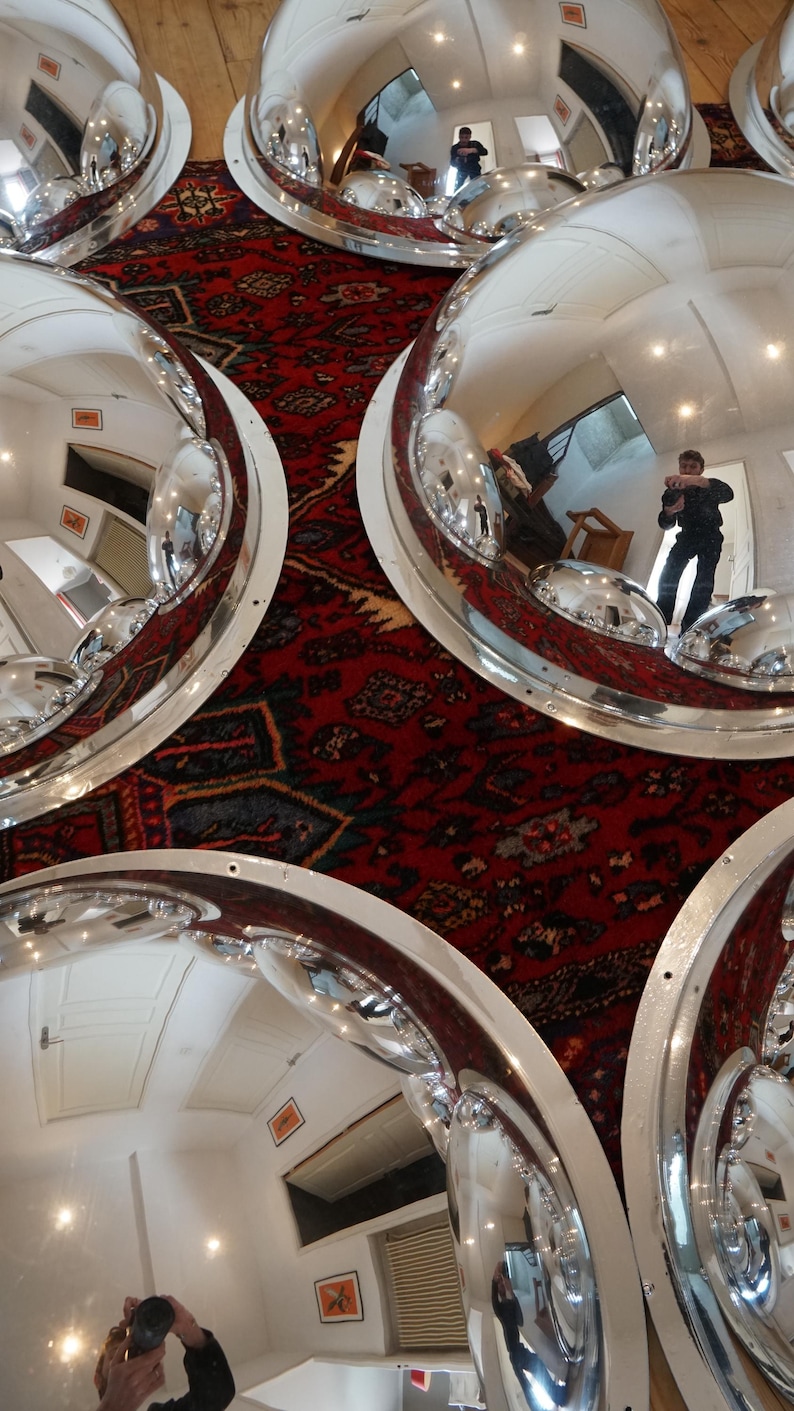 May include: Overhead view of multiple large, polished, chrome-like, hemispherical mirrors with white rims. The mirrors reflect the room and a red patterned rug. The mirrors are arranged in a circular pattern.