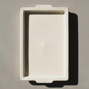Puede incluir: Una bandeja rectangular de cerámica en color blanco roto con una base plana y lisa y bordes elevados. La bandeja cuenta con dos pequeñas asas integradas en los lados cortos, ofreciendo un diseño limpio y funcional para servir u organizar objetos.