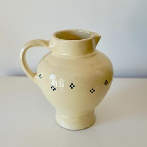 Puede incluir: Jarra de cerámica de color crema con cuerpo redondeado, asa y pico. La jarra está decorada con pequeños grupos de puntos negros espaciados. Es un objeto decorativo.