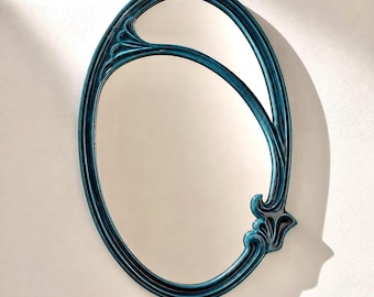 May include: An oval mirror featuring a deep teal, glossy frame with a raised, curved accent that divides the upper portion of the glass. The frame has a smooth, moulded appearance with a decorative, stylised floral-like detail on the lower right side.