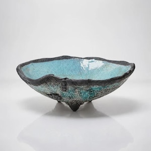 May include: A shallow, tripod ceramic bowl featuring a textured, dark charcoal-colored exterior with a grid-like pattern and small circular accents. The interior is finished with a glossy, crackled turquoise glaze that contrasts with the dark rim.