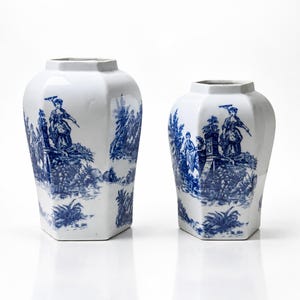 Può includere: Due vasi in ceramica bianca di forma esagonale, caratterizzati da un motivo blu in stile toile che raffigura figure in un paesaggio. I vasi presentano una finitura liscia e lucida e un'ampia apertura superiore, con un design coerente su entrambi.