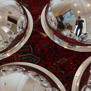 May include: Several large, polished chrome dome mirrors with white trim are arranged on a patterned red rug. The mirrors reflect the room and the person taking the photo.