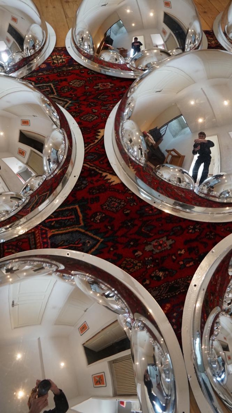 May include: Several large, polished chrome dome mirrors with white trim are arranged on a patterned red rug. The mirrors reflect the room and the person taking the photo.