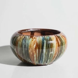 May include: A round ceramic bowl featuring a glossy glaze with vertical drip patterns in shades of brown, green, and blue. The interior of the bowl is a solid terracotta color, and the exterior surface displays a smooth, reflective finish.