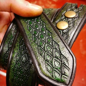 May include: A close-up of a green leather belt with a detailed embossed pattern. The belt has two brass buckles.