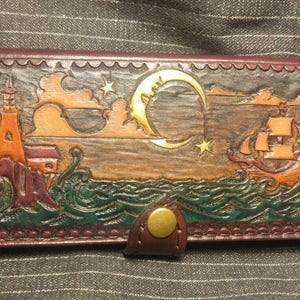 May include: A brown leather wallet with a tooled design of a lighthouse, a ship, and a crescent moon with stars. The wallet has a gold metal snap closure.