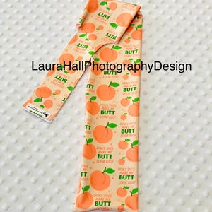 May include: A peach-colored fabric with a repeating pattern of peaches and the text "DOES THIS MAKE MY BUTT LOOK BIG?". The fabric is folded and displayed on a white, textured surface.
