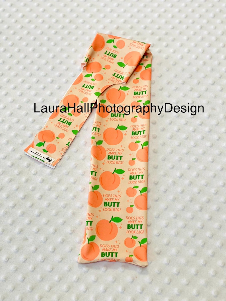 May include: A peach-colored fabric with a repeating pattern of peaches and the text "DOES THIS MAKE MY BUTT LOOK BIG?". The fabric is folded and displayed on a white, textured surface.