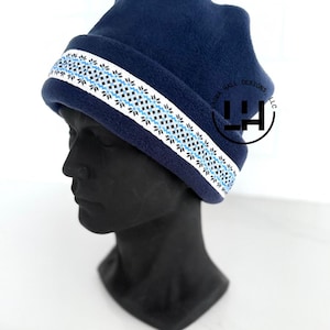 May include: A navy blue fleece beanie hat with a white band featuring a geometric pattern in black and light blue. The hat is displayed on a black mannequin head. The text "LAURA HALL DESIGNS, LLC" is visible on the side.