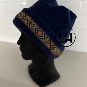 May include: A navy blue beanie featuring a decorative band with a multi-colored geometric pattern. The hat is displayed on a black mannequin head and includes a circular logo with the text Laura Hall Designs, LLC printed on the side.