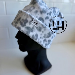May include: A grey and white leopard print beanie with a folded cuff. A small white rectangular tag with a black circular logo is sewn onto the cuff. The text LAURA HALL DESIGNS, LLC is printed in a circle around a black logo on the right side.
