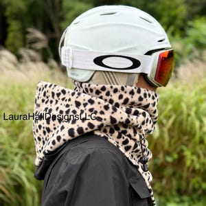 May include: A person wearing a light green ski helmet with red-tinted goggles and a leopard print neck warmer. The neck warmer is beige with black spots. The person is wearing a black jacket. The goggles are branded with the Oakley logo.
