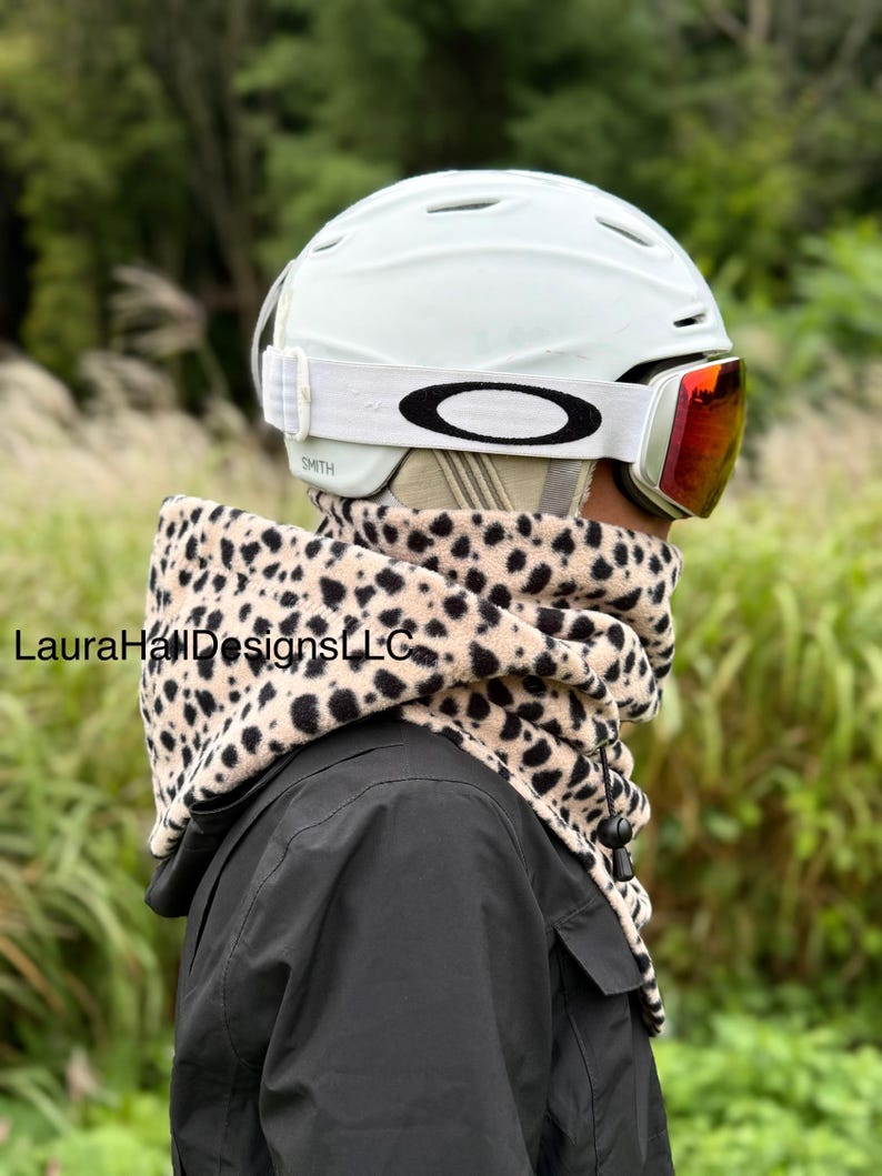 May include: A person wearing a light green ski helmet with red-tinted goggles and a leopard print neck warmer. The neck warmer is beige with black spots. The person is wearing a black jacket. The goggles are branded with the Oakley logo.