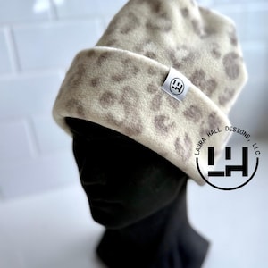 May include: A cream-colored beanie with a brown leopard print pattern. The hat is displayed on a black mannequin head. A small white tag with a logo is sewn on the front. The hat is made by Laura Hall Designs, LLC.