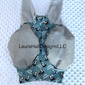 May include: A teal horse fly mask with grey mesh ear covers and eye protection. The fabric features a repeating pattern of black and white skunks with the text Little Stinker. The center of the mask displays the text LauraHallDesignsLLC.