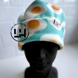 May include: A turquoise and white beanie hat with a repeating pattern of fried eggs. The hat is made of a soft material and is displayed on a black mannequin head. The hat has a small tag with the text "LH" on it.