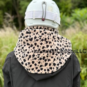 May include: A person wearing a light green Smith helmet with white straps and a tan and black spotted fleece neck warmer. The neck warmer is over a black jacket. The text "LauraHallDesignsLLC" is visible on the neck warmer.