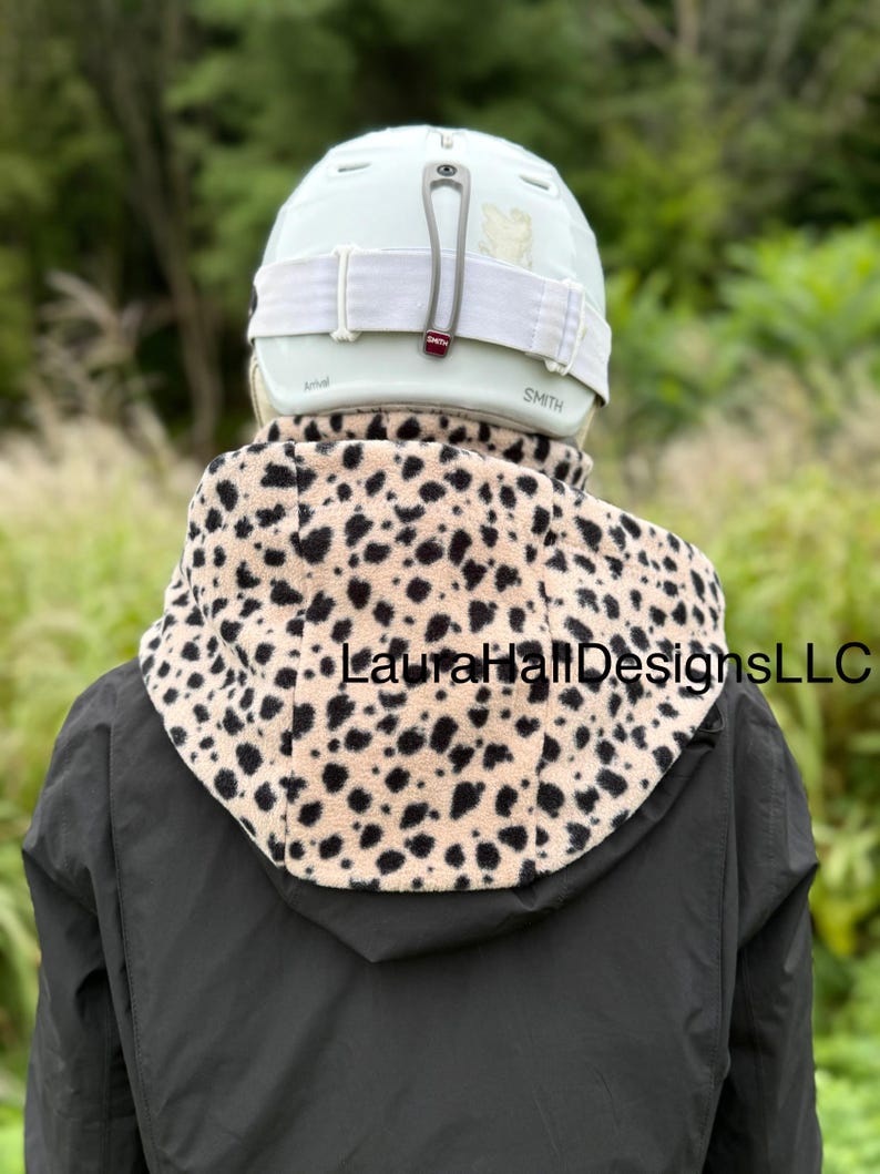 May include: A person wearing a light green Smith helmet with white straps and a tan and black spotted fleece neck warmer. The neck warmer is over a black jacket. The text "LauraHallDesignsLLC" is visible on the neck warmer.