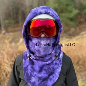 May include: A purple tie-dye fleece balaclava with a drawstring closure. The balaclava covers the head and neck, and is worn with a white helmet and red-tinted ski goggles. The text "LauraHallDesignsLLC" is printed on the balaclava.