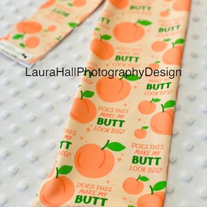 May include: Peach-themed leggings with a repeating pattern of peaches and the text "Does this make my butt look big?" in green. The leggings are peach-colored with a white background.