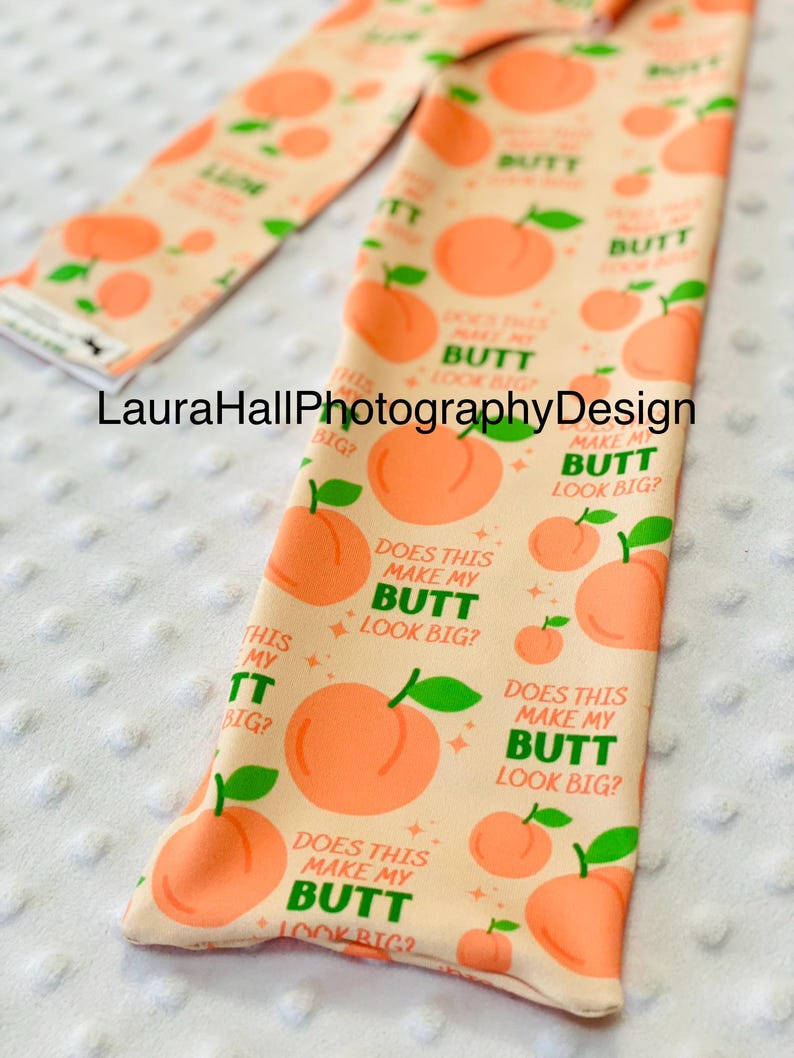 May include: Peach-themed leggings with a repeating pattern of peaches and the text "Does this make my butt look big?" in green. The leggings are peach-colored with a white background.