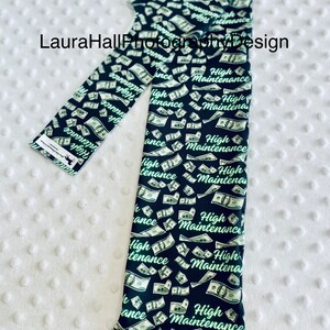 May include: Black leggings with a repeating pattern of falling dollar bills and the words "High Maintenance" in a green script. The leggings are folded on a white, textured surface.