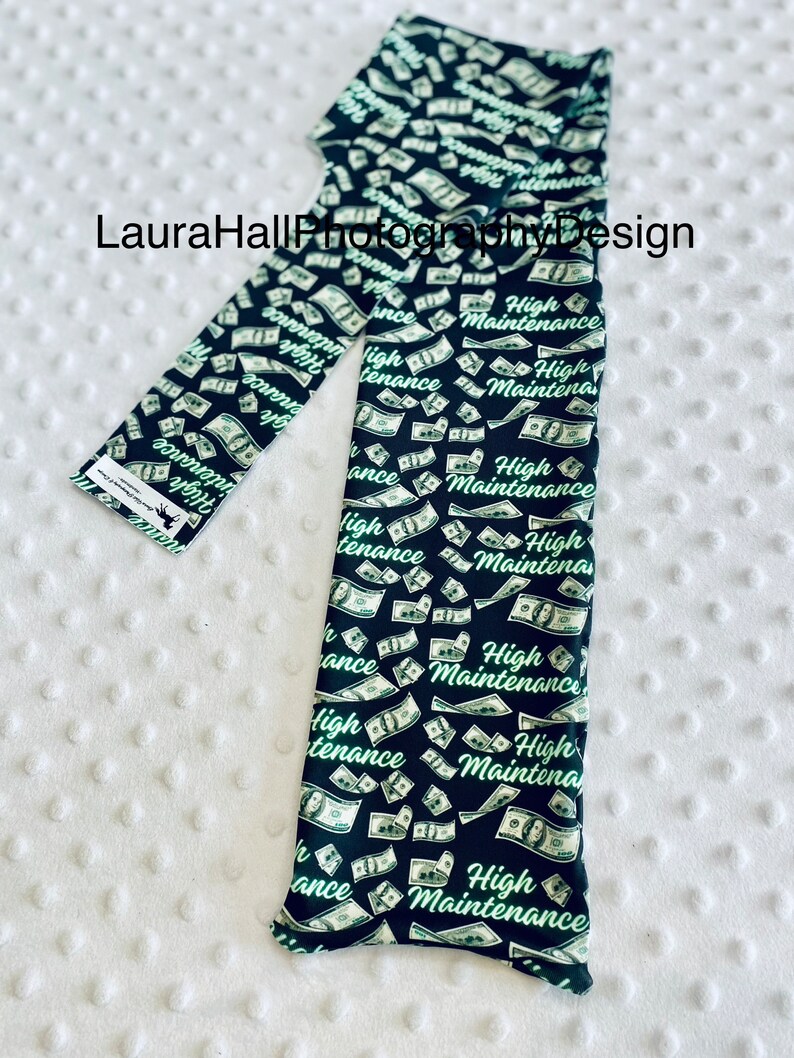 May include: Black leggings with a repeating pattern of falling dollar bills and the words "High Maintenance" in a green script. The leggings are folded on a white, textured surface.