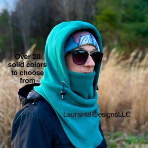 May include: A teal fleece hood and neck warmer with adjustable toggles. The text "Over 20 solid colors to choose from" is visible. The product is from LauraHallDesignsLLC. The person is wearing sunglasses and a patterned headband.