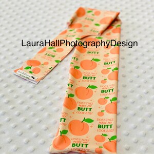 May include: Peach-colored fabric with a repeating pattern of peaches and the text "Does this make my butt look big?". The fabric is folded and appears to be a decorative item.