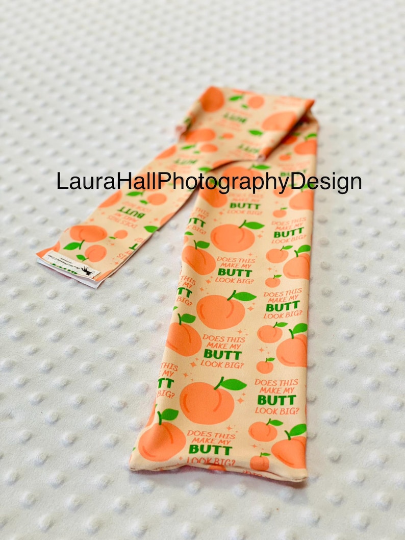 May include: Peach-colored fabric with a repeating pattern of peaches and the text "Does this make my butt look big?". The fabric is folded and appears to be a decorative item.