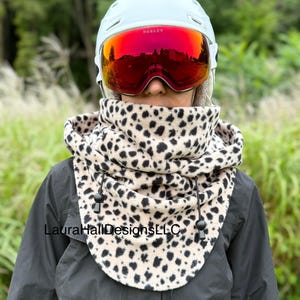 May include: A person wearing a light blue helmet with red Oakley goggles and a beige and black leopard print neck warmer. The neck warmer has a drawstring closure. The text "LauraHallDesignsLLC" is visible.