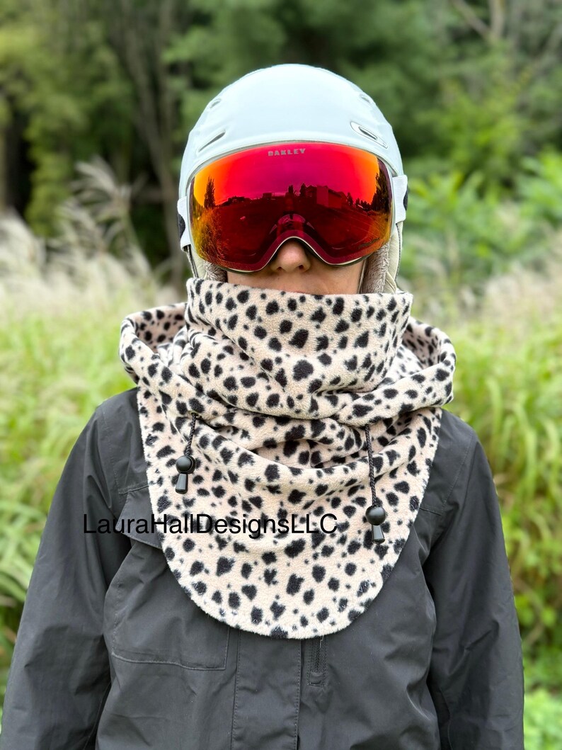 May include: A person wearing a light blue helmet with red Oakley goggles and a beige and black leopard print neck warmer. The neck warmer has a drawstring closure. The text "LauraHallDesignsLLC" is visible.