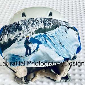 May include: A white ski helmet with a blue and white patterned cover featuring a skier on a snowy mountain. The cover has the text "LauraHallPhotographyDesign" printed on it.