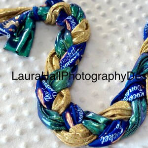 May include: A braided hair tie made of blue, green, and gold fabric with the words "Rocket's Journey" printed on it.
