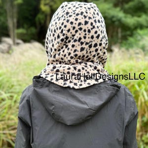 May include: A hooded scarf with a tan and black animal print. The scarf is worn over a dark gray jacket. The text "Laura Hall Designs LLC" is printed on the scarf.