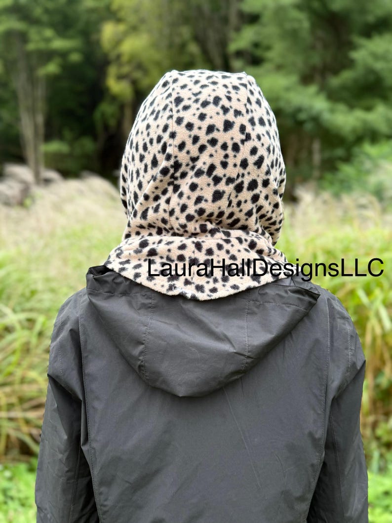 May include: A hooded scarf with a tan and black animal print. The scarf is worn over a dark gray jacket. The text "Laura Hall Designs LLC" is printed on the scarf.
