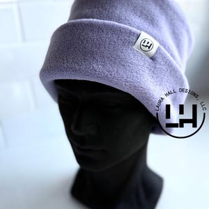 May include: A light purple fleece beanie with a folded cuff. A small white rectangular tag is sewn onto the cuff, featuring a black logo with the letters LH. The beanie is displayed on a black mannequin head, and a circular watermark reads LAURA HALL DESIGNS, LLC.