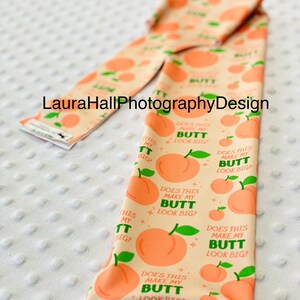 May include: A peach-colored fabric item with a repeating pattern of peaches and the text "DOES THIS MAKE MY BUTT LOOK BIG?". The item also has the text "THIS MY BUTT BIG?" and "BUTT".