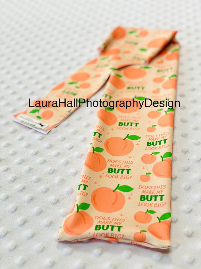 May include: A peach-colored fabric item with a repeating pattern of peaches and the text "DOES THIS MAKE MY BUTT LOOK BIG?". The item also has the text "THIS MY BUTT BIG?" and "BUTT".