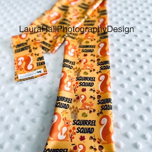 May include: A yellow fabric headband with a repeating pattern of squirrels, acorns, and mushrooms. The text "Squirrel Squad" is repeated throughout the pattern.