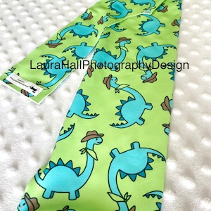 Könnte beinhalten: Ein grüner Stoff mit einem sich wiederholenden Muster aus blauen Dinosauriern, die Cowboyhüte tragen. Der Stoff ist in der Mitte gefaltet und der Text "LauraHallPhotographyDesign" ist sichtbar.