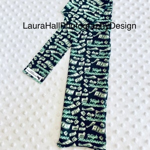 May include: Black leggings with a repeating pattern of falling dollar bills and the words "High Maintenance" in a green script. The leggings are displayed on a white, textured surface.