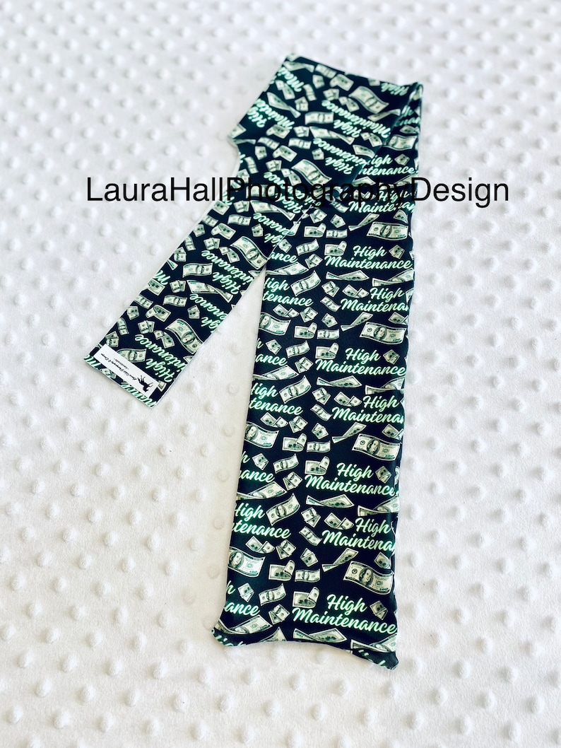 May include: Black leggings with a repeating pattern of falling dollar bills and the words "High Maintenance" in a green script. The leggings are displayed on a white, textured surface.